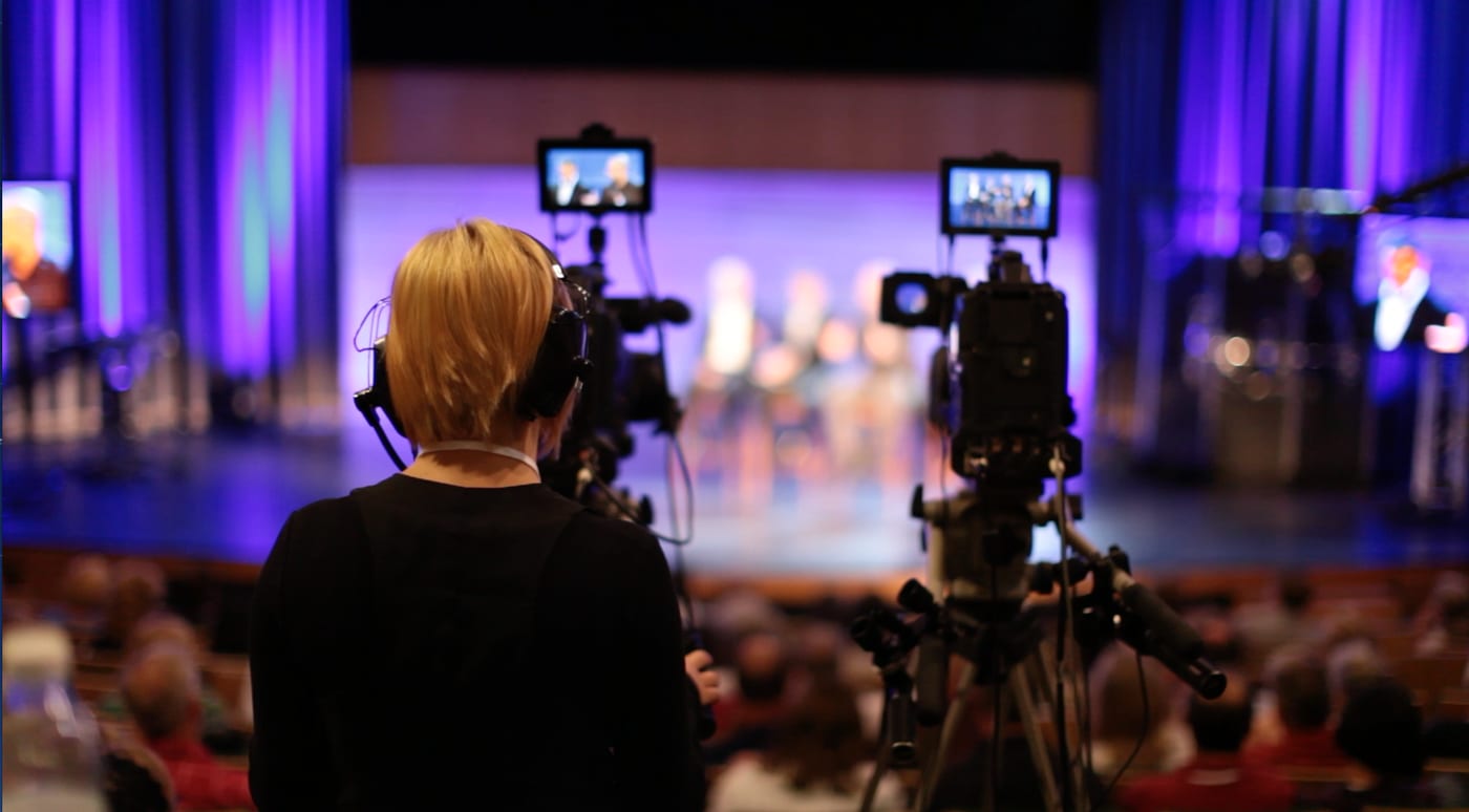 A broadcast camera operator wearing a headset at a live event, framing a stage shot on the camera monitor.