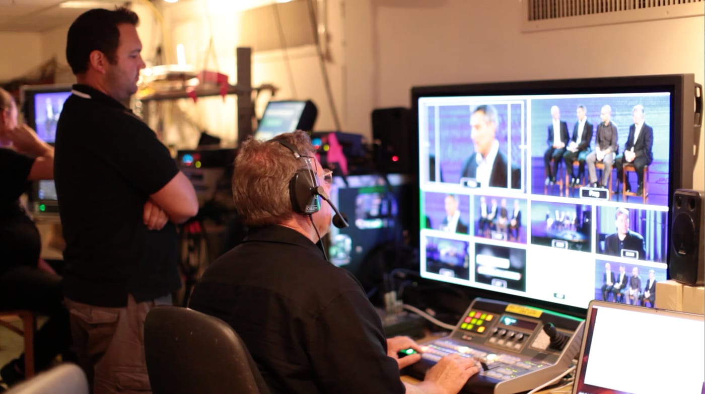 The Zoom Presence Pro CEO at the switcher in a live broadcast control room, wearing a headset and directing the show, with a wall of camera monitors in front of him.