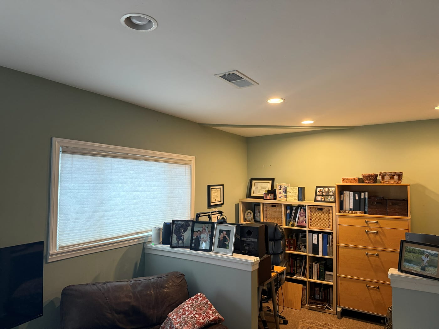 A wide photo of a home office: green walls, a large window with the blind down, a leather sofa, and bookshelves along the back wall.