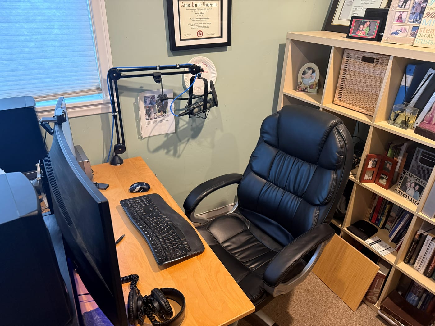 A close photo of the desk where calls happen: a boom microphone arm, monitors, a keyboard, a large leather chair, and a crowded bookcase beside it.