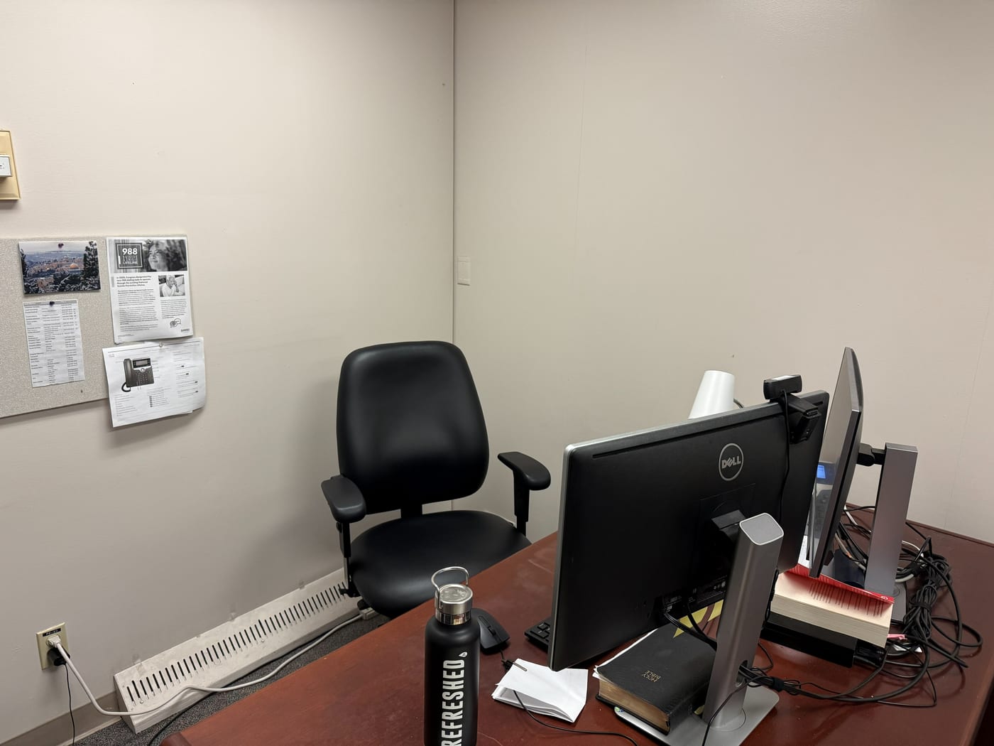 A close photo of the corner where video calls happen: an office chair against a blank wall, a bulletin board of papers, and a webcam clipped to a monitor.