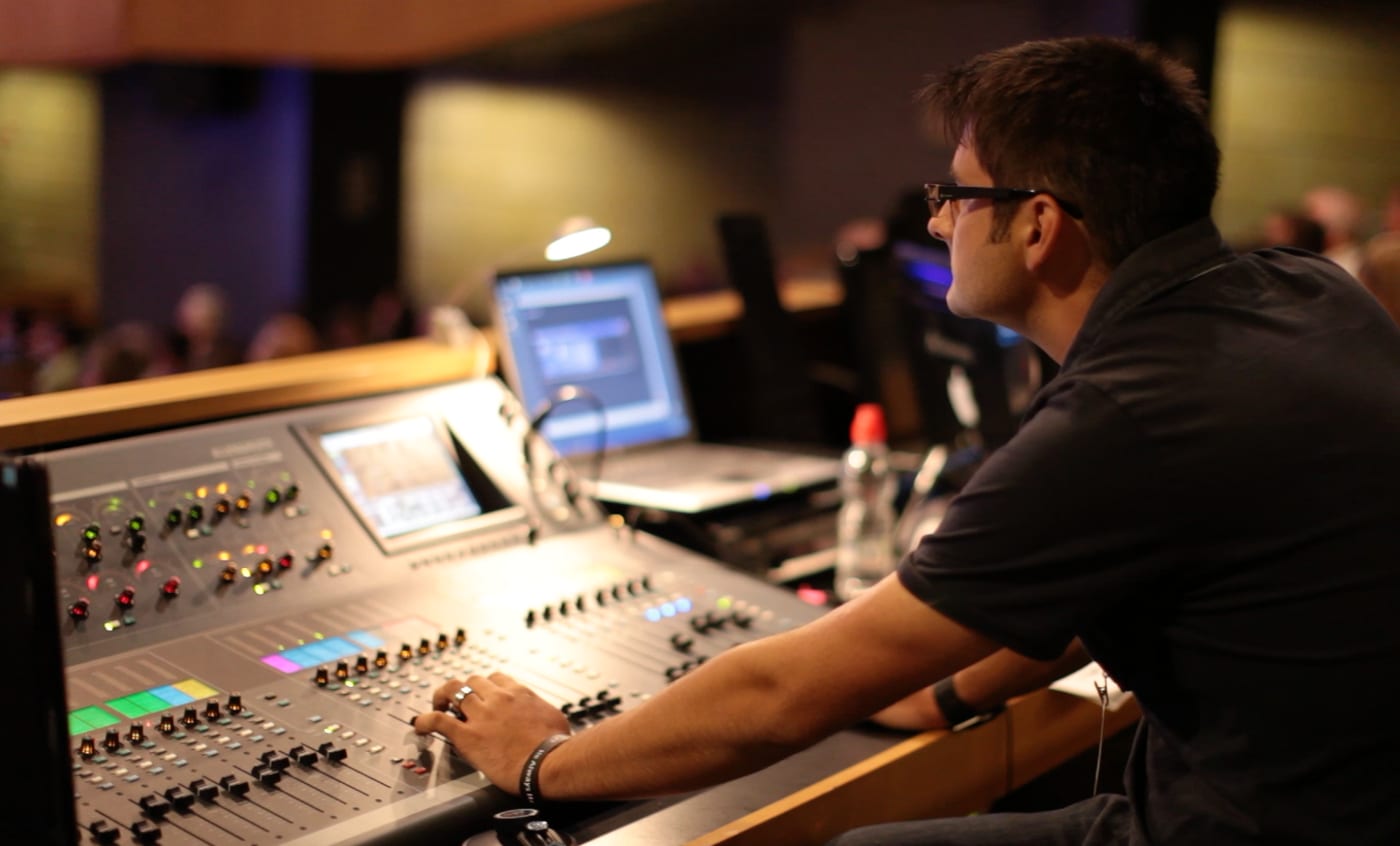 A sound engineer at a mixing console during a live event, with the audience and stage behind him.